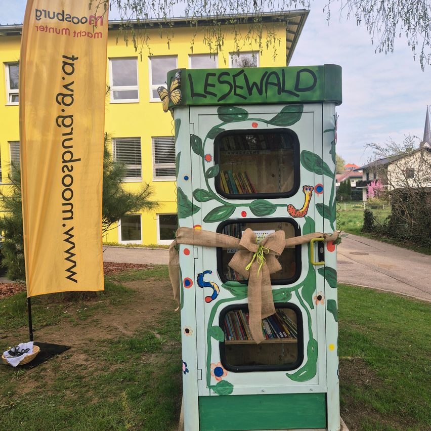 Eine bunte Büchertauschbox mit dem Wort Lesewald ist mit Pflanzen und einer Schleife verziert. Links steht ein gelber Banner.