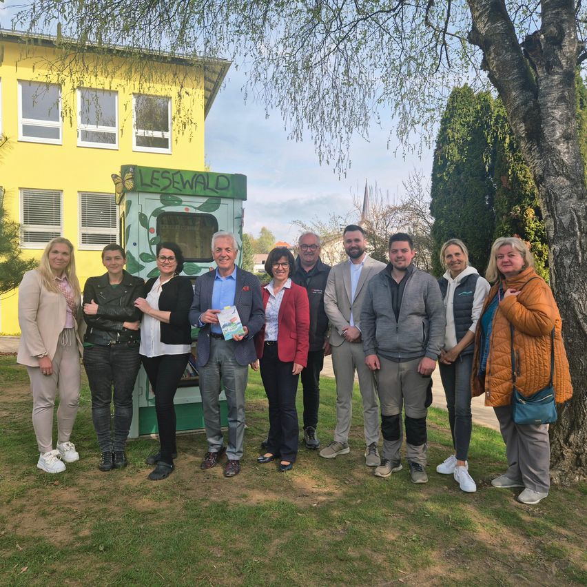 Eine Gruppe von Menschen, darunter einige Frauen und Männer, posiert für ein Foto vor einem gelben Gebäude und einem grünen Kiosk mit der Aufschrift Lesewald.