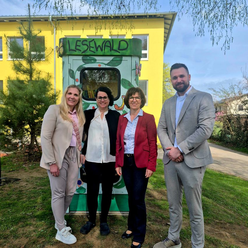 Vier Personen stehen vor einem hellgelben Gebäude mit der Aufschrift Lesewld. Sie scheinen für ein Foto zu posieren. Dahinter befinden sich Bäume und ein Weg.