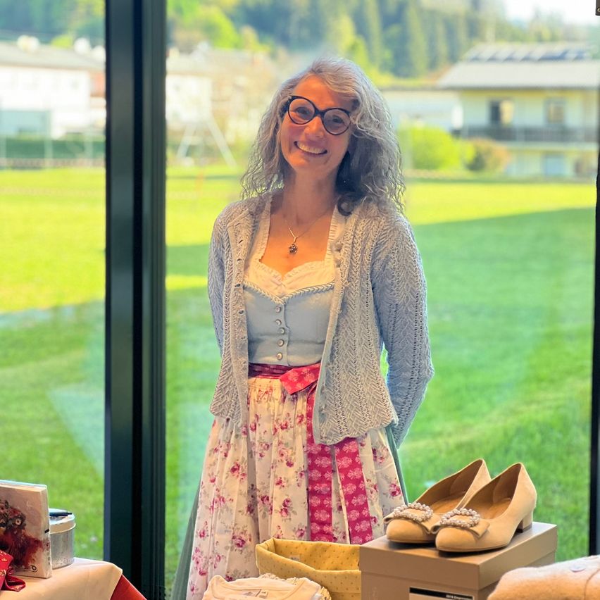 Eine Frau mit grauem Haar und Brille steht lächelnd vor einer Glaswand. Hinter ihr befindet sich ein grünes Feld und ein Haus. Vor ihr stehen Gegenstände wie Schuhe, eine Box und ein Buch.