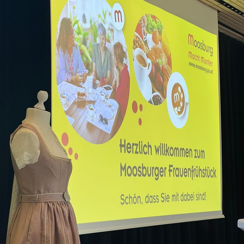 Eine Präsentation mit einem Kleidermannequin auf der Bühne, die eine gelbe Leinwand mit einer Willkommensnachricht für das Moosburger Frauenfrühstück zeigt. Bilder einer Familienmahlzeit und einer Kaffeetasse sind zu sehen.