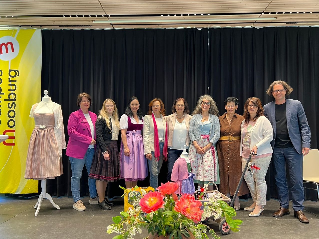 Eine Gruppe von Frauen in verschiedenen Outfits steht vor einem schwarzen Vorhang, mit Mannequins und Blumen im Vordergrund.