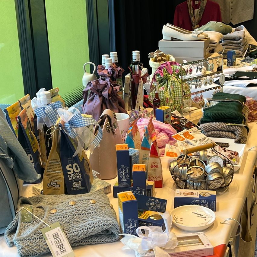 Ein Tisch mit verschiedenen Artikeln wie einem Korb, Boxen, einem Teller und einer Flasche. Ein grauer gestrickter Pullover mit einem Anhänger ist ebenfalls vorhanden. Der Tisch ist mit einem weißen Tuch bedeckt.