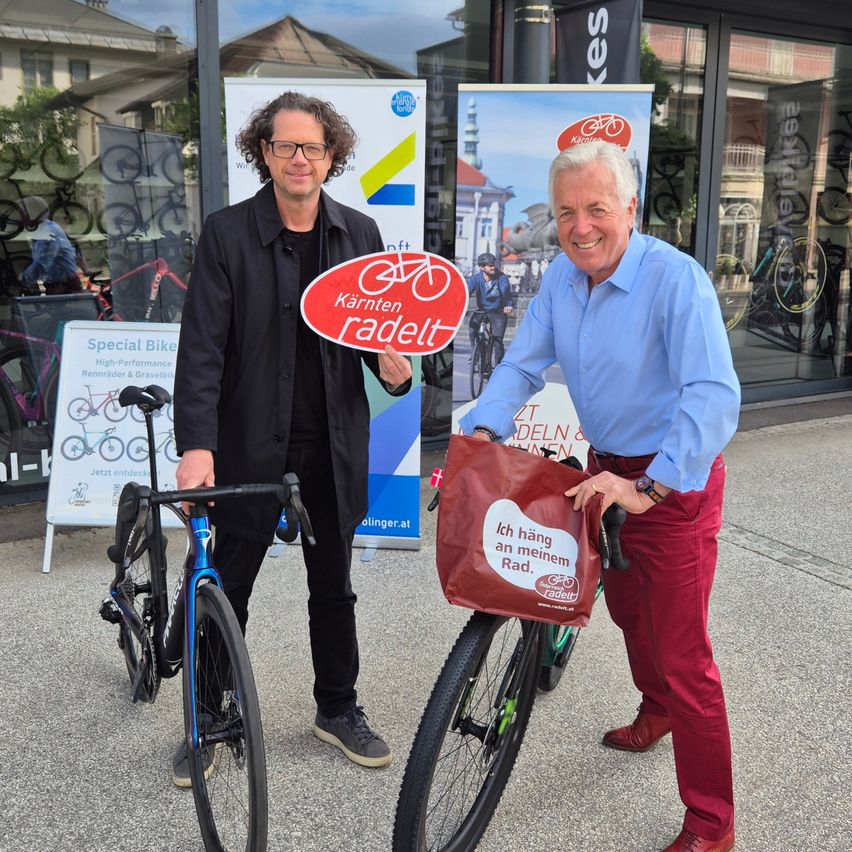Zwei Männer posieren mit Fahrrädern vor einem Geschäft, einer hält ein Schild 'Kärnten radelt' und der andere trägt eine rote Tasche mit dem weißen Text 'Ich hänge an meinem Rad'.