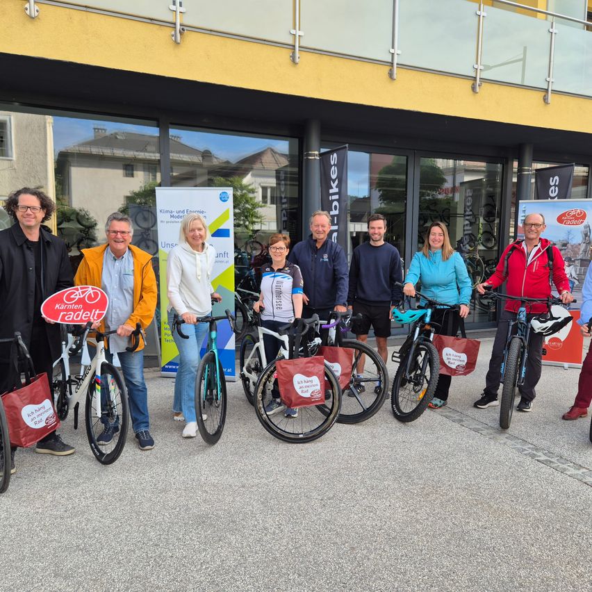 Eine Gruppe von Menschen in Winterkleidung steht neben Fahrrädern vor einem Gebäude mit Glaswänden. Sie halten Banner in den Händen und lächeln.
