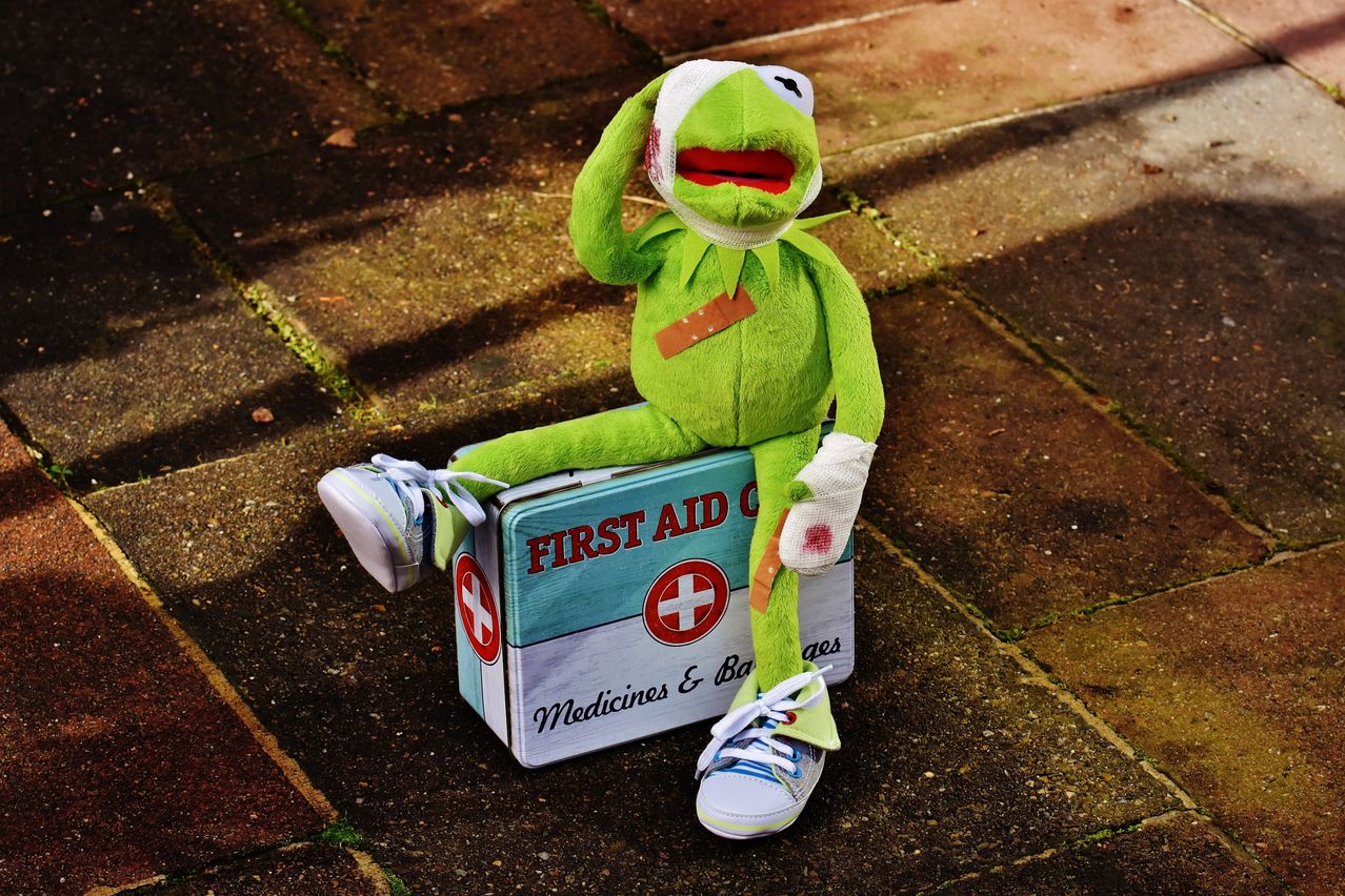 Ein Plüsch-Kermit der Frosch sitzt auf einer Erste-Hilfe-Kiste. Er hat Verbände am Kopf und am Arm.