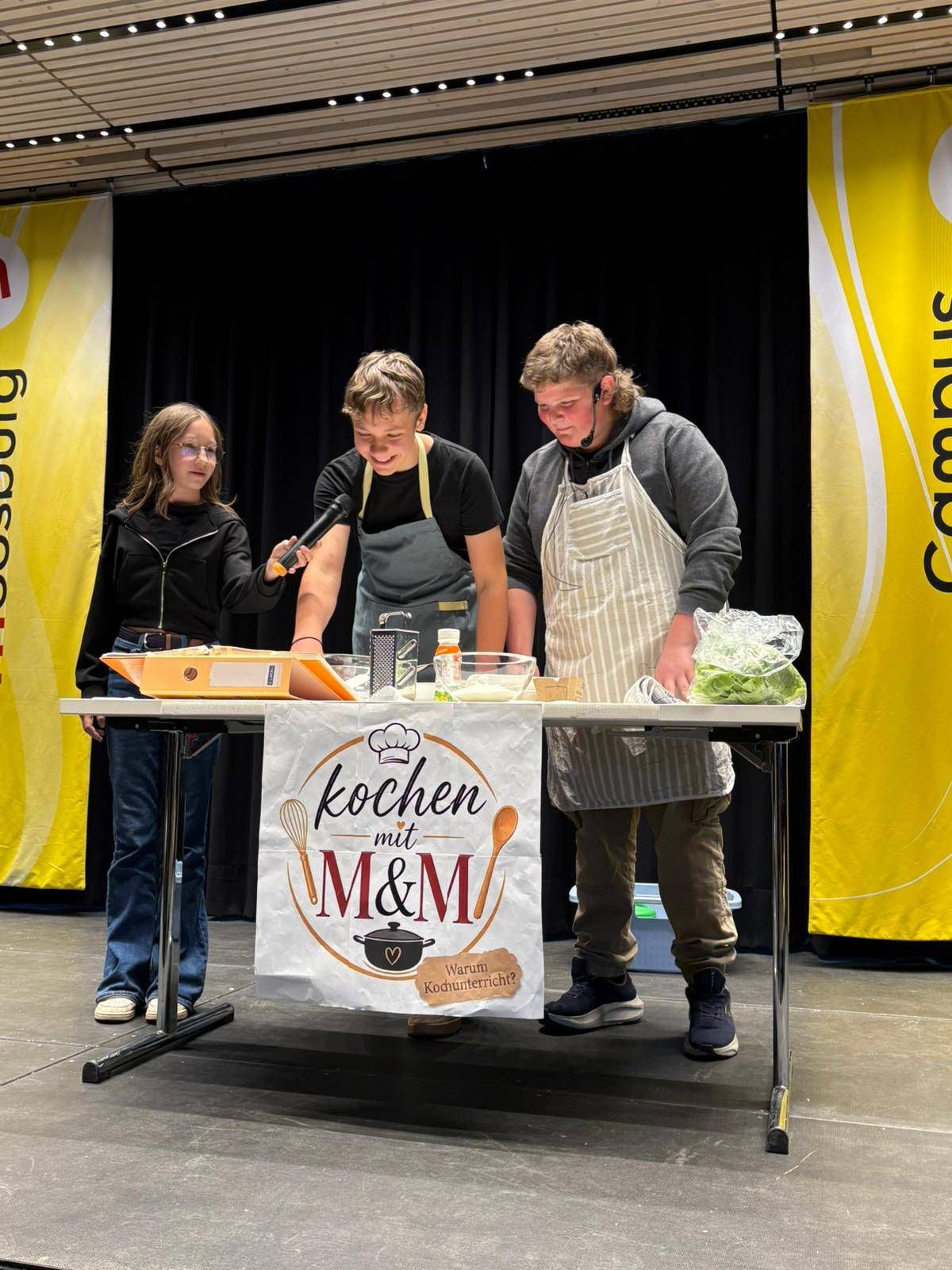 Drei Personen stehen hinter einem Tisch mit einem Banner, auf dem 'kochen mit M&M' steht. Sie bereiten Essen mit einer Frau vor, die ein Mikrofon hält. Der Tisch ist mit Zutaten beladen. Hinter ihnen befinden sich Banner mit Logos.