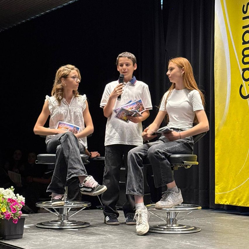 Drei Schüler sitzen auf der Bühne, einer hält ein Mikrofon, während andere Bücher halten. Ein Blumentopf ist in der Nähe. Die Bühne hat einen gelben Hintergrund.