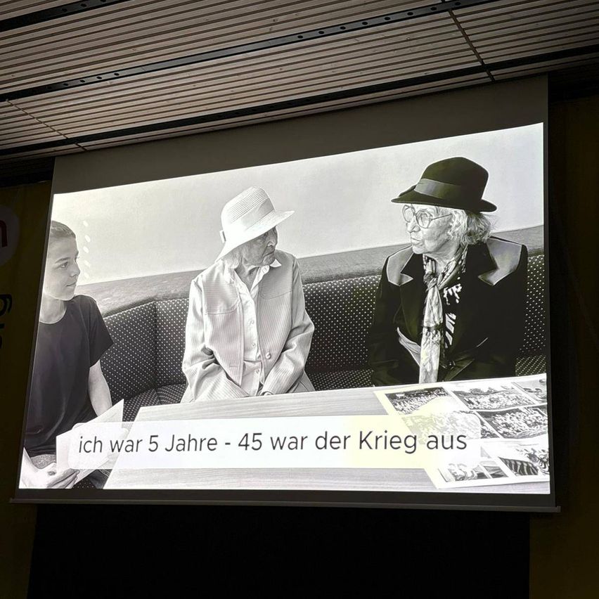 Ein Schwarz-Weiß-Foto auf einer Projektionsleinwand zeigt zwei ältere Frauen, die mit einem jungen Mann auf einer Bank sitzen. Eine Frau trägt einen Hut und einen Schal, die andere trägt eine Brille und einen Mantel. Der Text lautet: 'Ich war 5 Jahre alt - 45 Jahre ist der Krieg vorbei.'