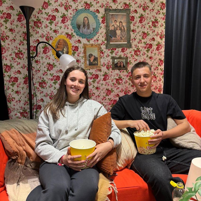 Zwei Personen sitzen auf einem Sofa und halten Popcorn in gelben Eimern. Hinter ihnen schmückt eine Blumentapete die Wand mit mehreren Familienfotos.
