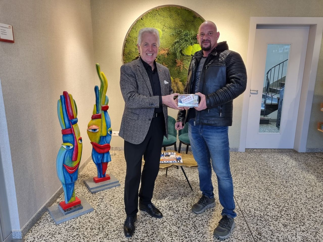 Zwei Männer stehen in einem Innenbereich mit einem lächelnden Gesicht. Eine Person auf der linken Seite hält eine Box und eine andere Person hält ein Buch auf dem Tisch.