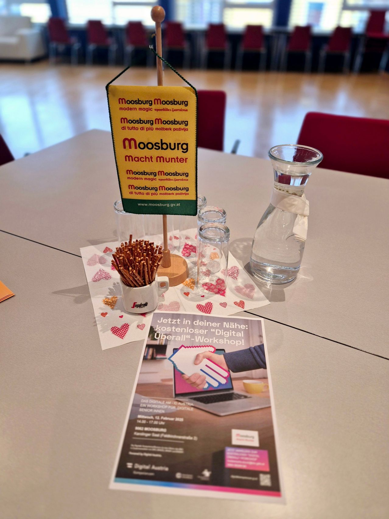 Ein Tisch mit einem Schild, das 'Moosburg Moosburg macht Munter' besagt. Der Tisch hat einen Becher, ein Glas und eine Wasserkaraffe. Ein Flyer liegt auf dem Tisch mit einer Hand, die einen Laptop hält.