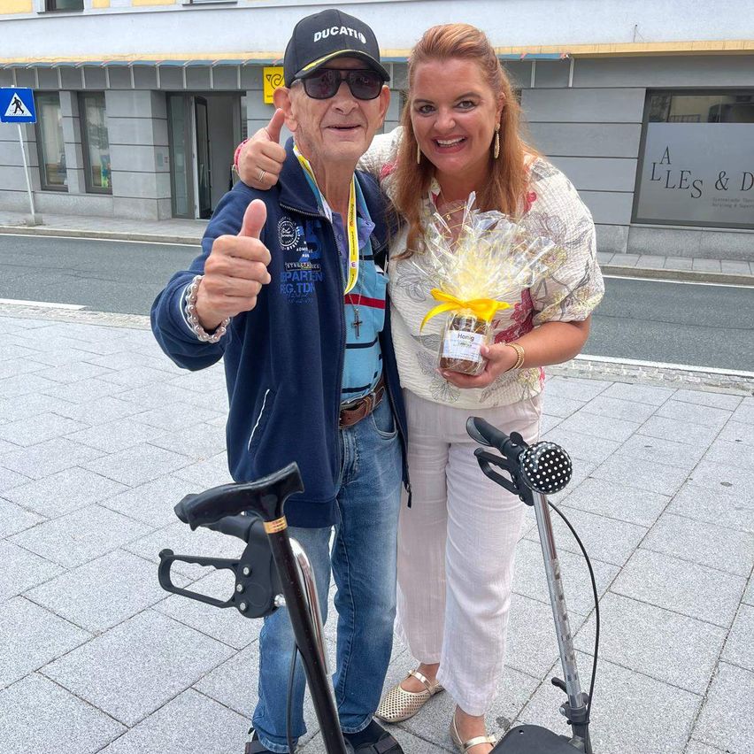 Ein älterer Mann mit Kappe und Sonnenbrille gibt ein Daumen hoch, während er neben einer Frau steht, die ein verpacktes Geschenk auf einem Stadtpfad hält.