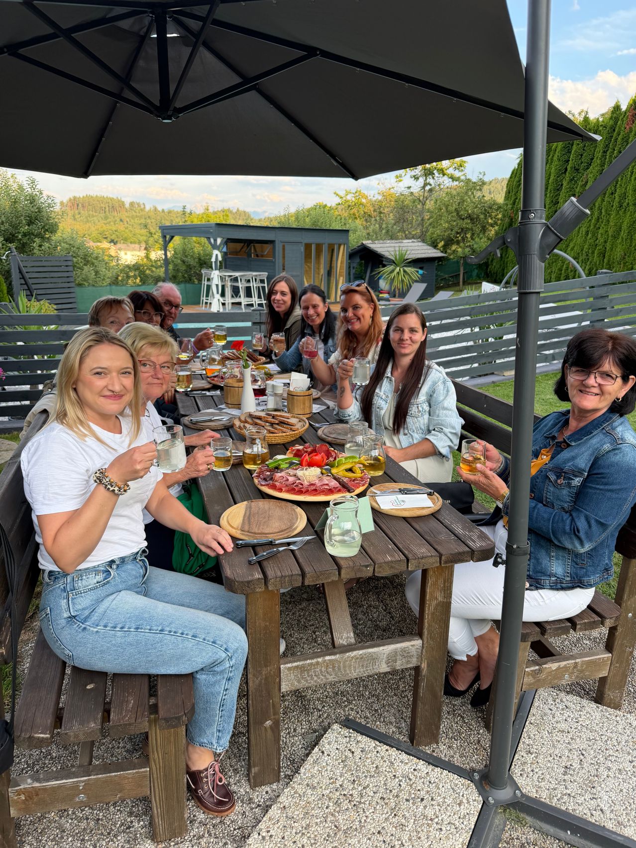Eine Gruppe von Frauen sitzt um einen Tisch im Freien, lächelt und hält Getränke in den Händen. Der Tisch ist mit Tellern mit Essen, Gläsern und einer Schüssel Obst gedeckt. Der Hintergrund zeigt eine Umzäunung und viel Grün.