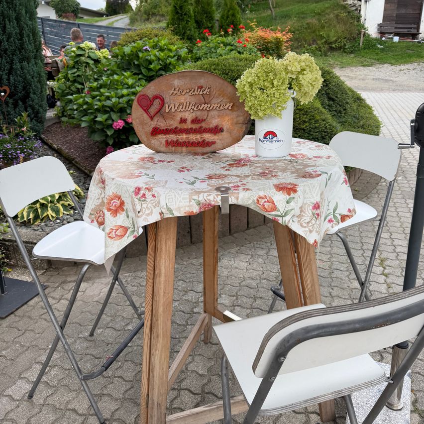 Ein hölzerner Tisch mit einer Tischdecke mit Blumenmuster und einem Holzschild, das Herzlich Willkommen in der Buchenwalder Wassermühle besagt. Eine weiße Vase mit grünen Blumen steht auf dem Tisch. Auf beiden Seiten des Tisches stehen zwei weiße Klappstühle.