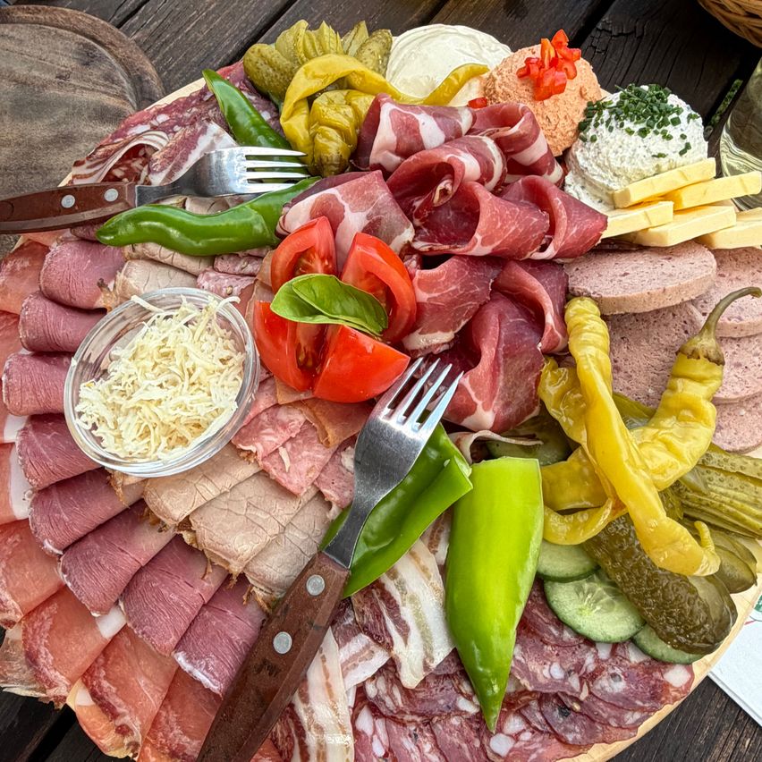 Ein Holztablett mit verschiedenen Wurstsorten, Käse, Tomaten, Paprika und Beilagen auf einem Holztisch.