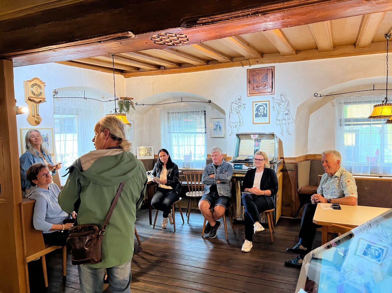 Eine Gruppe von Menschen sitzt in einem Raum mit Holzböden und weißen Wänden. Einige sitzen auf Stühlen, andere stehen. Eine Frau in einer grünen Jacke steht in der Nähe der Wand.