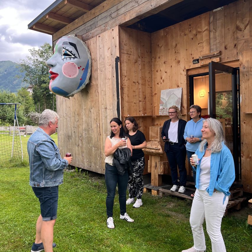 Eine Gruppe von Menschen steht vor einem Holzhaus mit einer großen bemalten Gesichtsmaske an der Wand und unterhält sich bei Getränken.