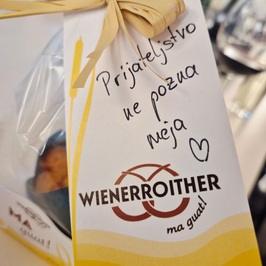 Ein Geschenkanhänger mit einer Schleife, der den Text 'Prijateljstvo ne pozna meja' und das Logo von 'Wienerroither' zeigt. Der Anhänger ist an einem Plastikbeutel befestigt, der ein Lebensmittel enthält.