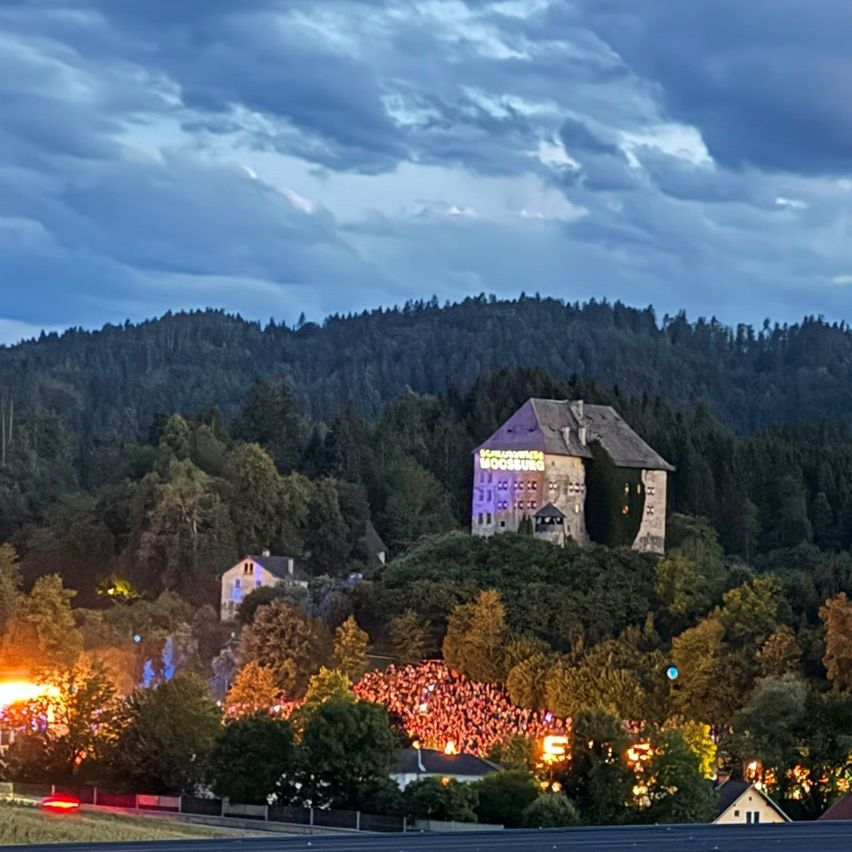 Eine Burg auf einem Hügel mit einer Menge unten, beleuchtet von Lichtern unter einem bewölkten Himmel.