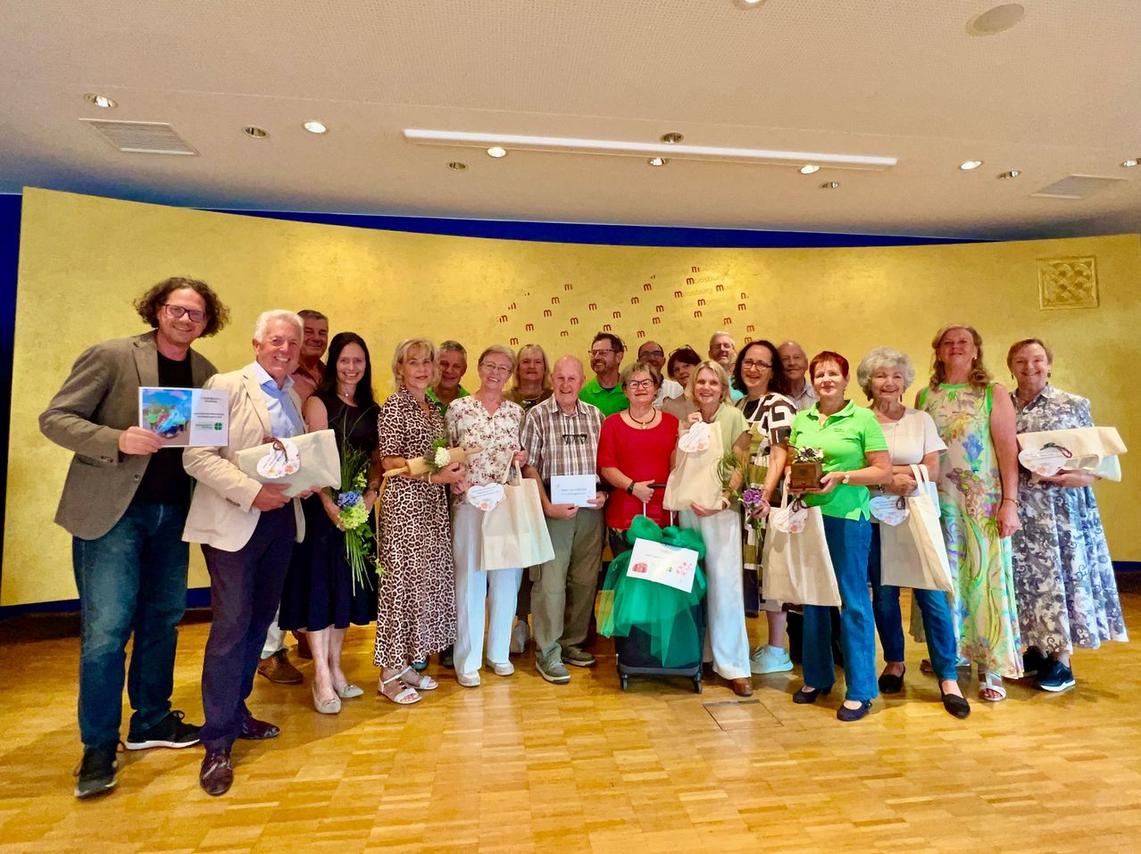 Eine Gruppe von Menschen steht in einem Raum, lächelt und hält Blumen, Taschen und Urkunden. Sie sind in verschiedenen Outfits gekleidet.