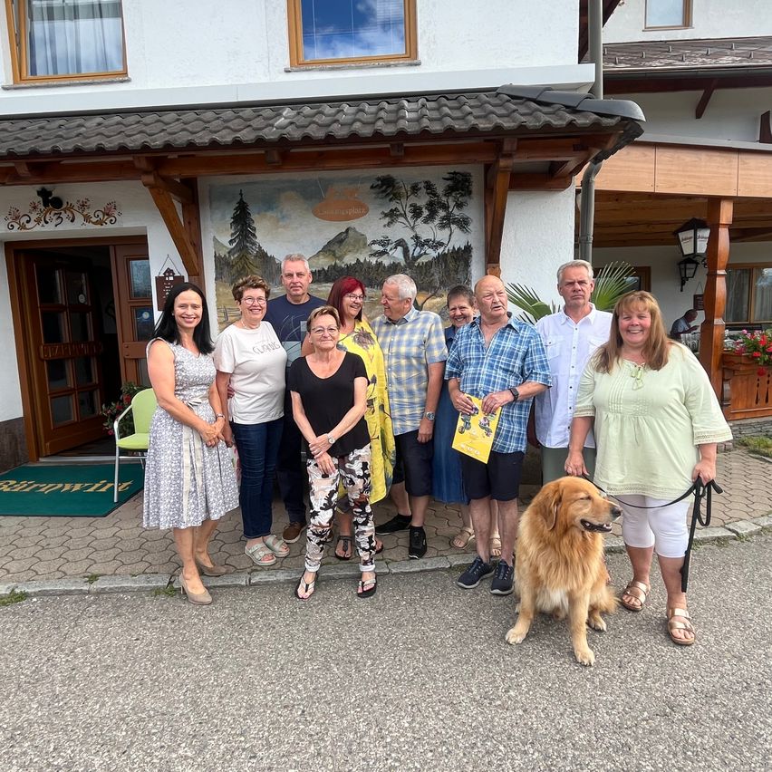 Eine Gruppe von Menschen, einschließlich eines Hundes, steht vor einem Gebäude mit einem Wandbild und einer Lampe. Sie lächeln und posieren für ein Foto.