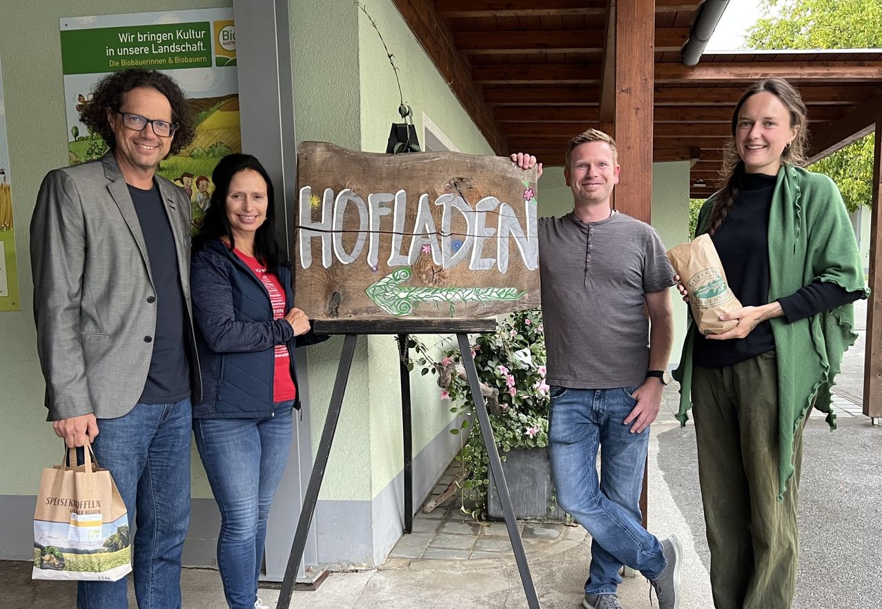Vier Personen stehen vor einem Holzschild, auf dem 'HOFLADEN' steht. Das Schild steht auf einem Ständer, und die Personen lächeln. Dahinter befindet sich eine Wand mit einem Poster und einem Topf mit Pflanzen.