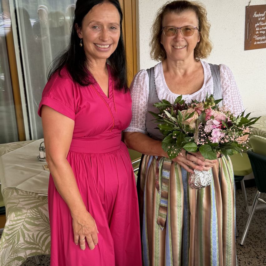 Zwei Frauen stehen nebeneinander, eine in einem rosa Kleid und die andere in einem traditionellen Kleid, halten einen Blumenstrauß. Sie lächeln und scheinen für ein Foto zu posieren. Hinter ihnen steht ein Tisch mit einer Tischdecke und ein Schild an der Wand.