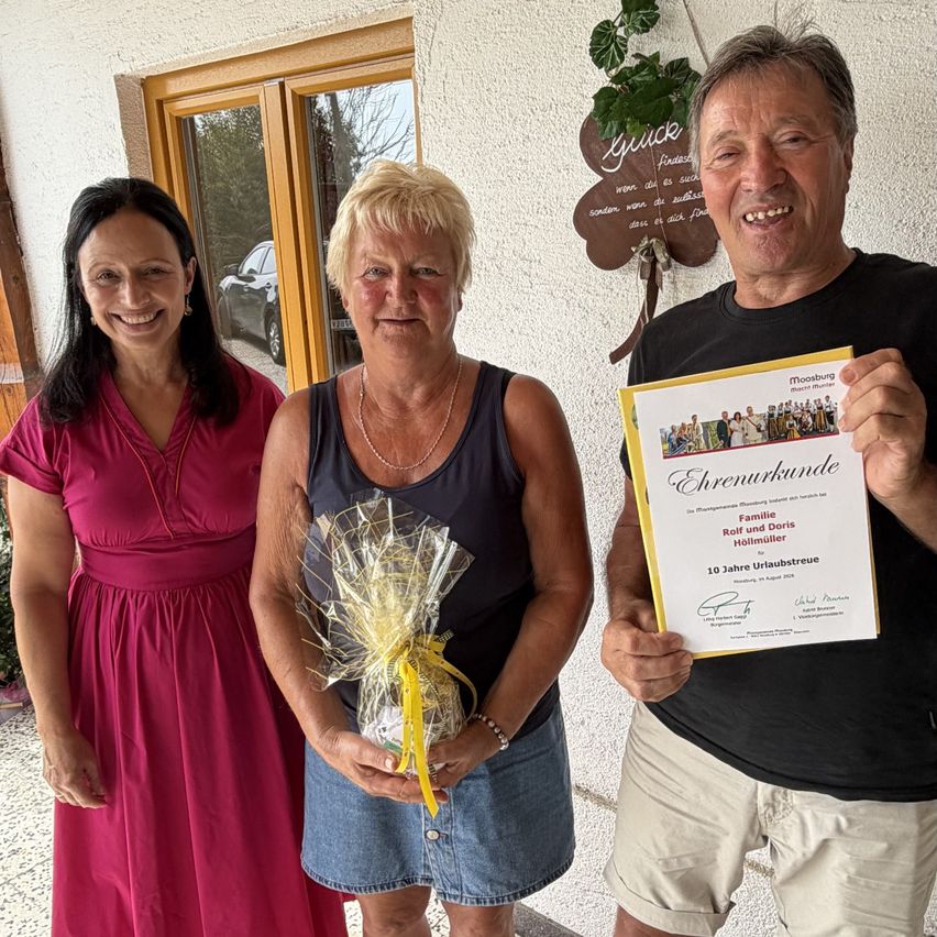 Drei Personen stehen lächelnd zusammen, einer hält ein Zertifikat und ein Geschenk. Die Frau links trägt ein rosa Kleid, die Frau in der Mitte ein ärmelloses Top und der Mann rechts ein schwarzes T-Shirt. Hinter ihnen ist eine Wand mit einem dekorativen Kleeblatt.