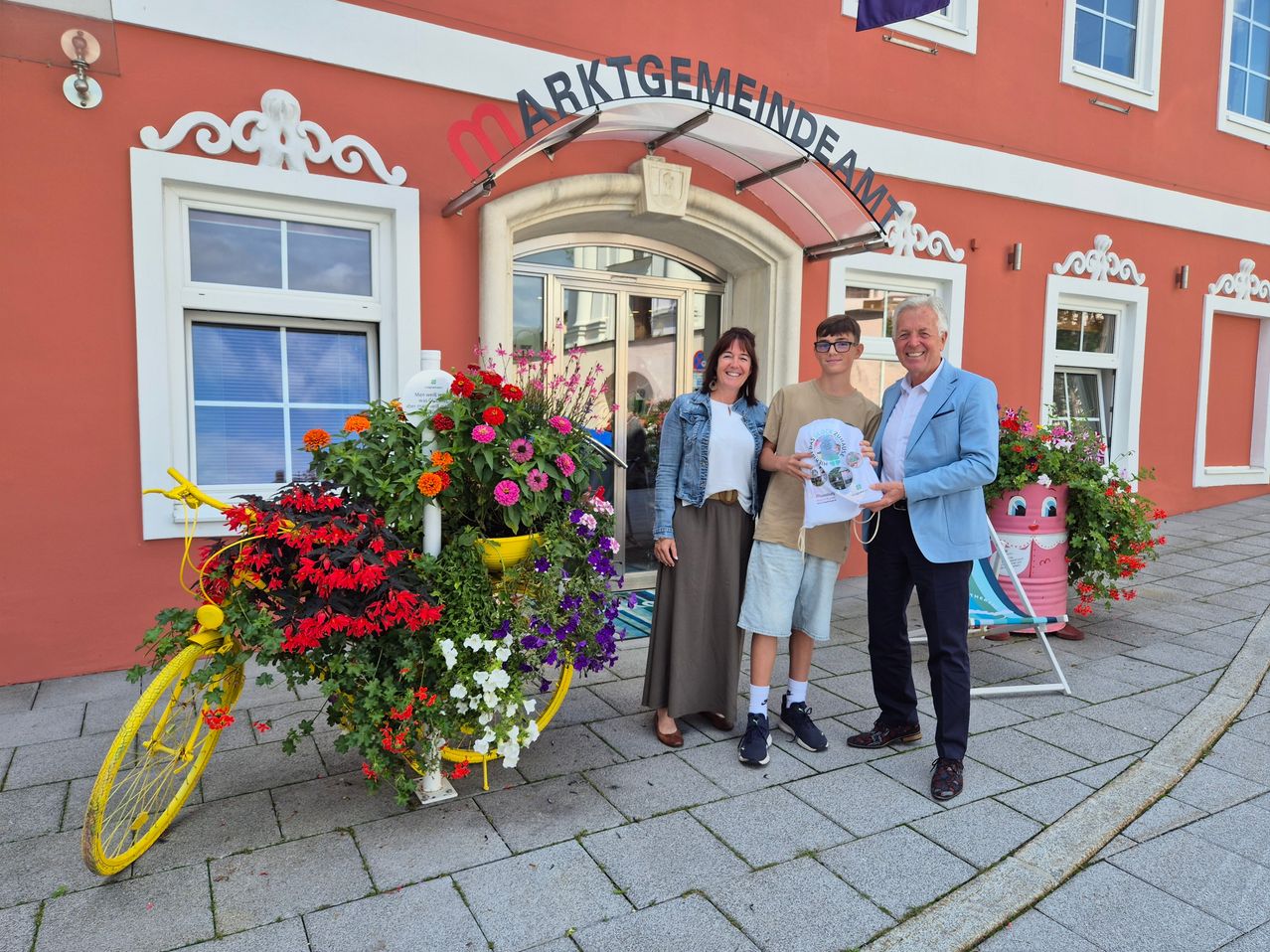 Drei Personen stehen vor einem Gebäude mit einem blumengeschmückten Fahrrad. Der Mann auf der rechten Seite hält ein Papier in der Hand und die Frau in der Mitte lächelt. Der Mann auf der linken Seite trägt eine Brille.