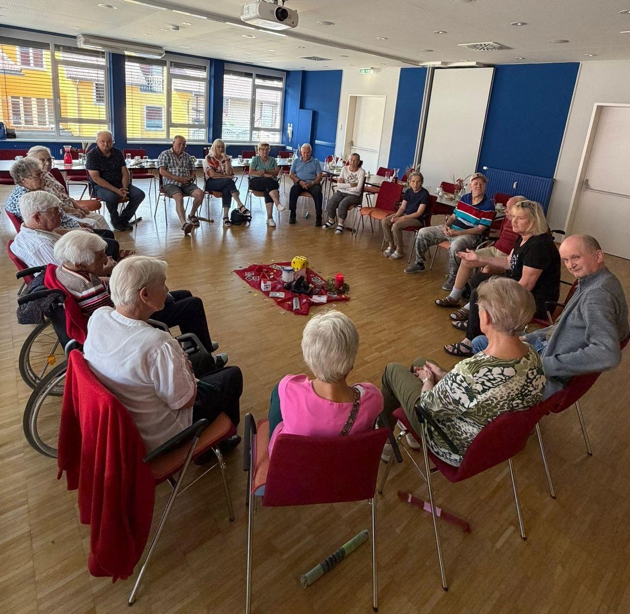 Eine Gruppe älterer Menschen sitzt im Kreis in einem Raum mit blauen Wänden und Holzböden. Einige sitzen in Rollstühlen. Ein Tisch in der Mitte hat verschiedene Gegenstände.