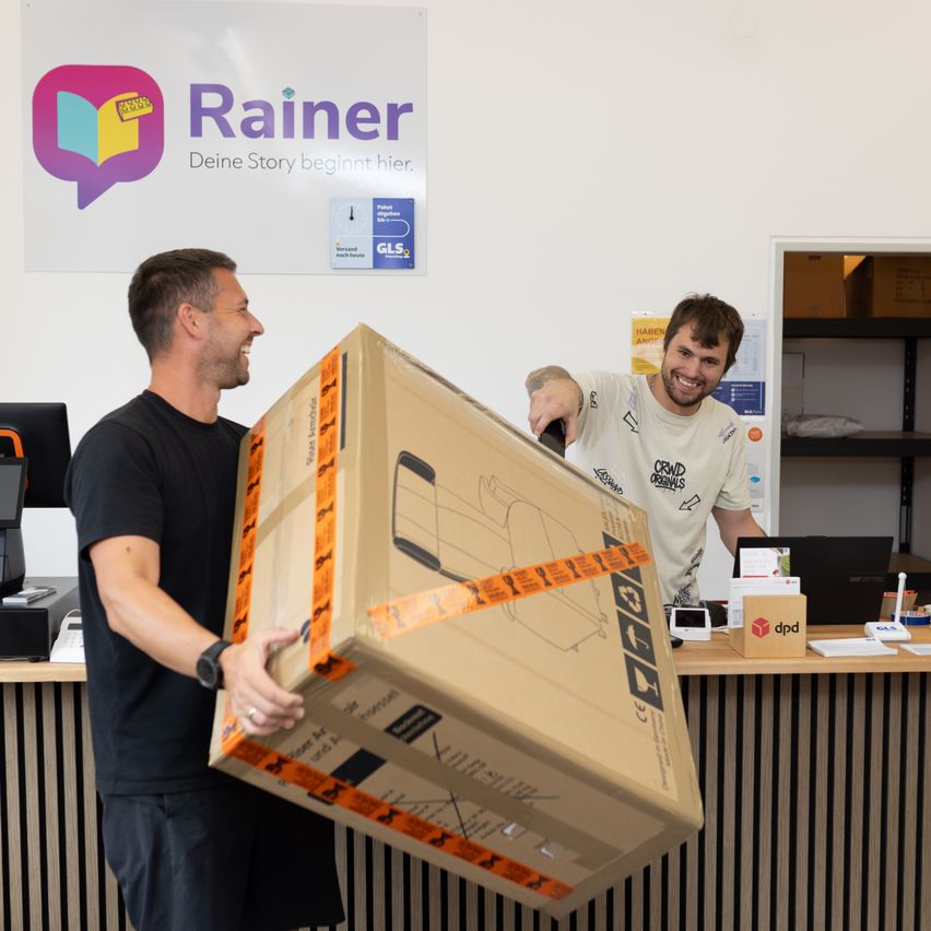 Zwei Männer tragen eine große Kiste in einem Büro. Ein Mann lächelt und der andere hält die Kiste. Hinter ihnen steht ein Tresen mit einem Laptop, einer Kiste und einem Schild.