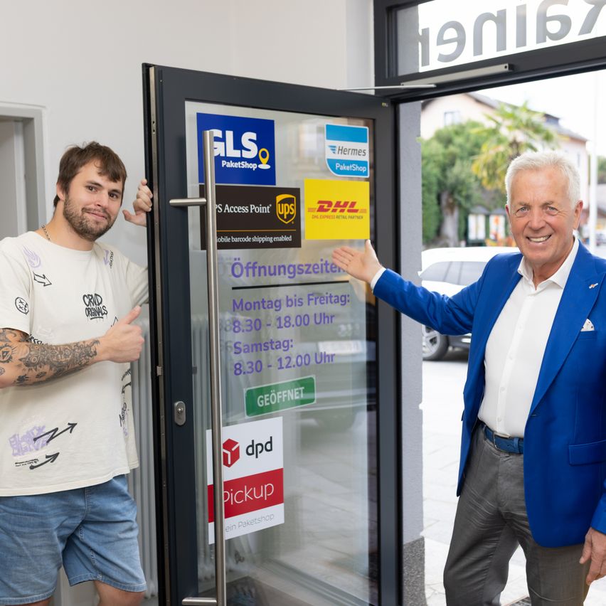 Zwei Männer stehen neben einer offenen Tür mit einem Schild, das 'Öffnungszeiten' anzeigt. Der Mann auf der linken Seite trägt ein weißes T-Shirt mit einer Tätowierung am Arm. Der Mann auf der rechten Seite trägt einen blauen Anzug und hält den Türgriff. Hinter ihnen ist ein Auto auf der Straße geparkt.