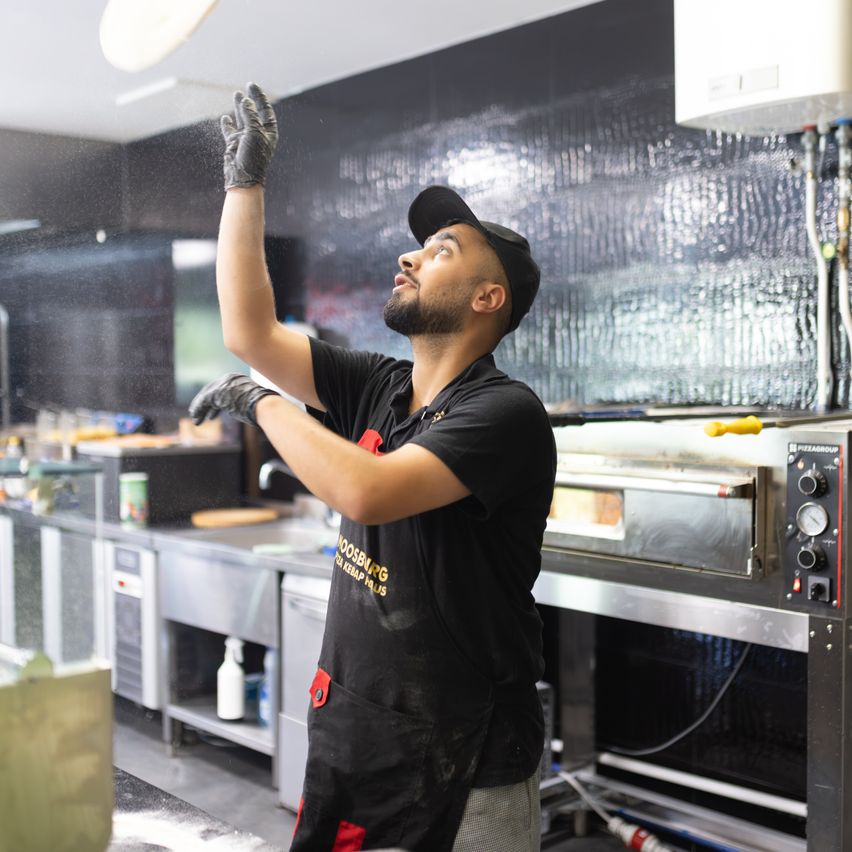 Ein Mann in einem schwarzen Apron und Hut arbeitet in einer Küche und streut etwas auf eine Pizza.