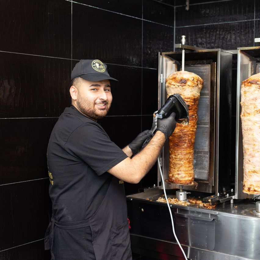 Ein Mann in einem schwarzen Shirt und Hut schneidet Fleisch von einem Drehspieß. Er steht vor einer schwarzen gefliesten Wand.