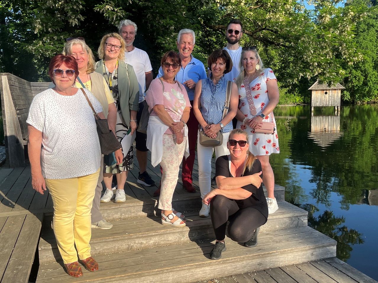 Eine Gruppe von Menschen, hauptsächlich Frauen, posiert für ein Foto auf einem Holzsteg am See, einige tragen Brillen und Sonnenbrillen. Bäume und ein kleines Haus sind im Hintergrund.
