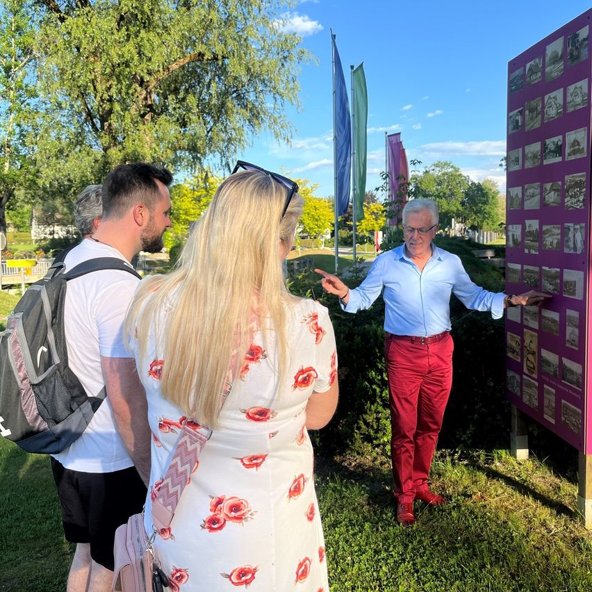 Ein Mann in einem blauen Hemd und roten Hosen erklärt etwas einer Frau mit einem Blumenkleid und einem anderen Mann mit einem Rucksack. Sie stehen in einem Park mit einem großen lila Poster.