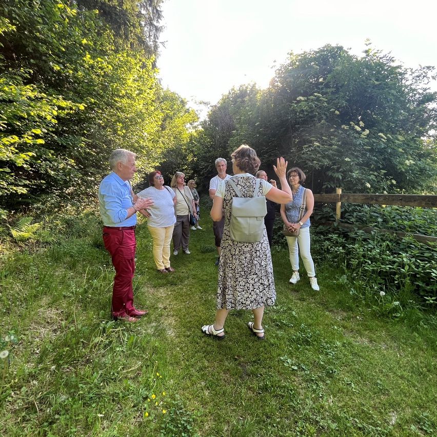 Eine Gruppe von Menschen geht in einem Grasbereich mit einem Holzzaun. Sie scheinen Touristen zu sein, eine Frau winkt. Die Umgebung ist im Freien mit Grün und Sonnenlicht.