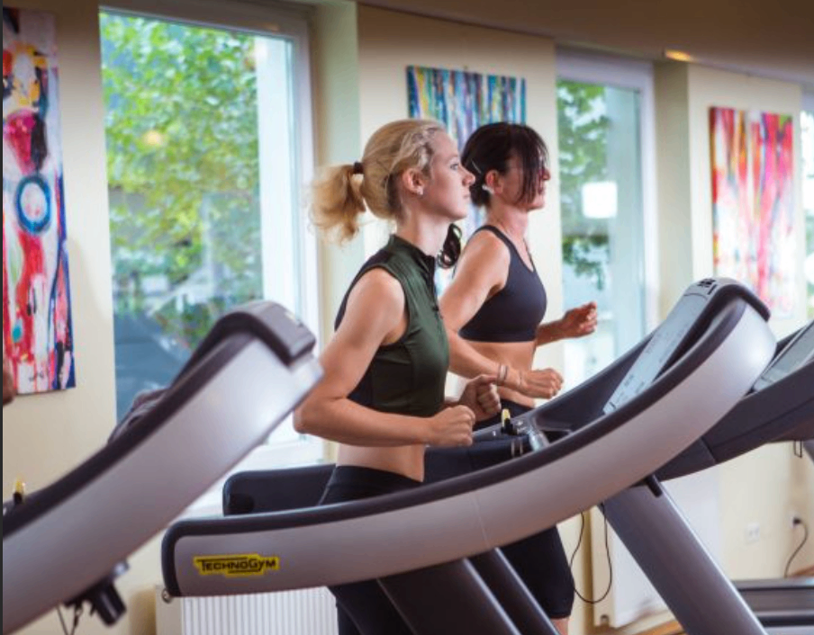 Zwei Frauen laufen auf Laufbändern in einem Fitnessstudio. Die Frau auf der linken Seite schaut nach vorne, während die Frau auf der rechten sie beobachtet. Eine Wand mit Fenstern und Kunst hängt hinter ihnen.