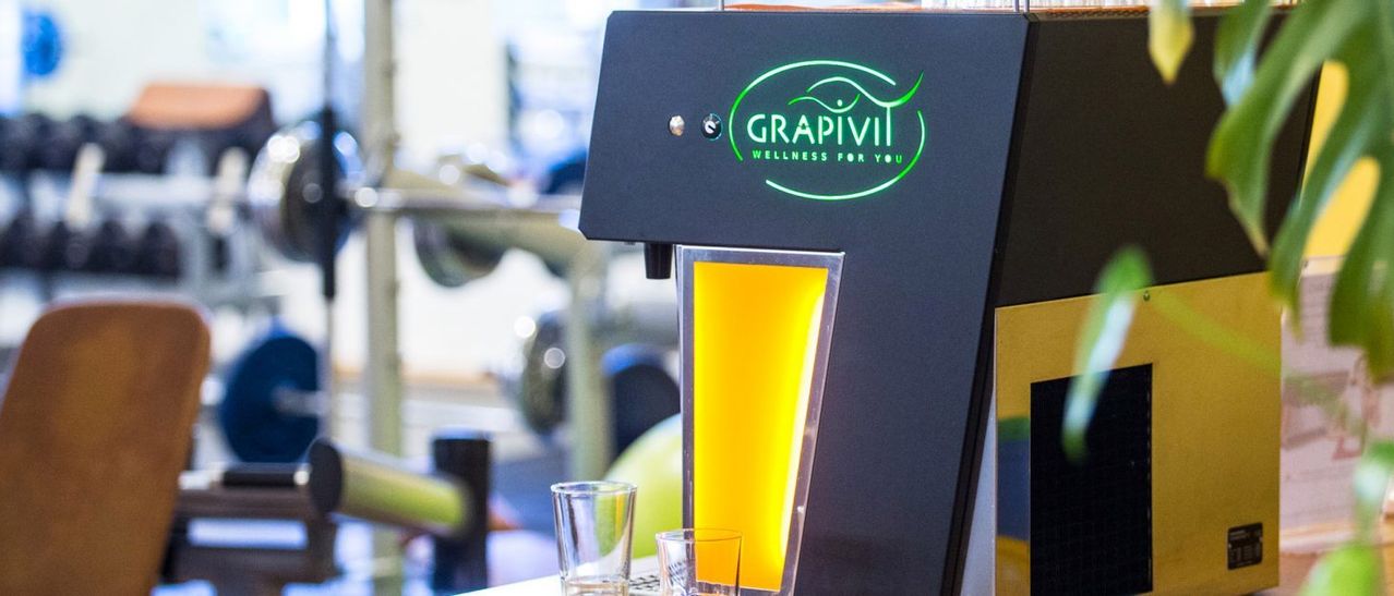 Eine Grapivii-Wellness-Maschine mit zwei Gläsern, gefüllt mit gelber Flüssigkeit, auf einem Tisch in einem Fitnessstudio.
