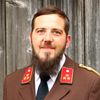 Ein Mann mit Bart und Schnurrbart trägt eine braune Uniform mit roten Sternen auf der Schulter und Namensschild 'INDERST' vor einem hölzernen Hintergrund.