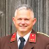 Ein lächelnder Mann in einem Militäruniform mit Medaillen und einem Namensschild 'MAUHART'. Er steht vor einer Holzwand.