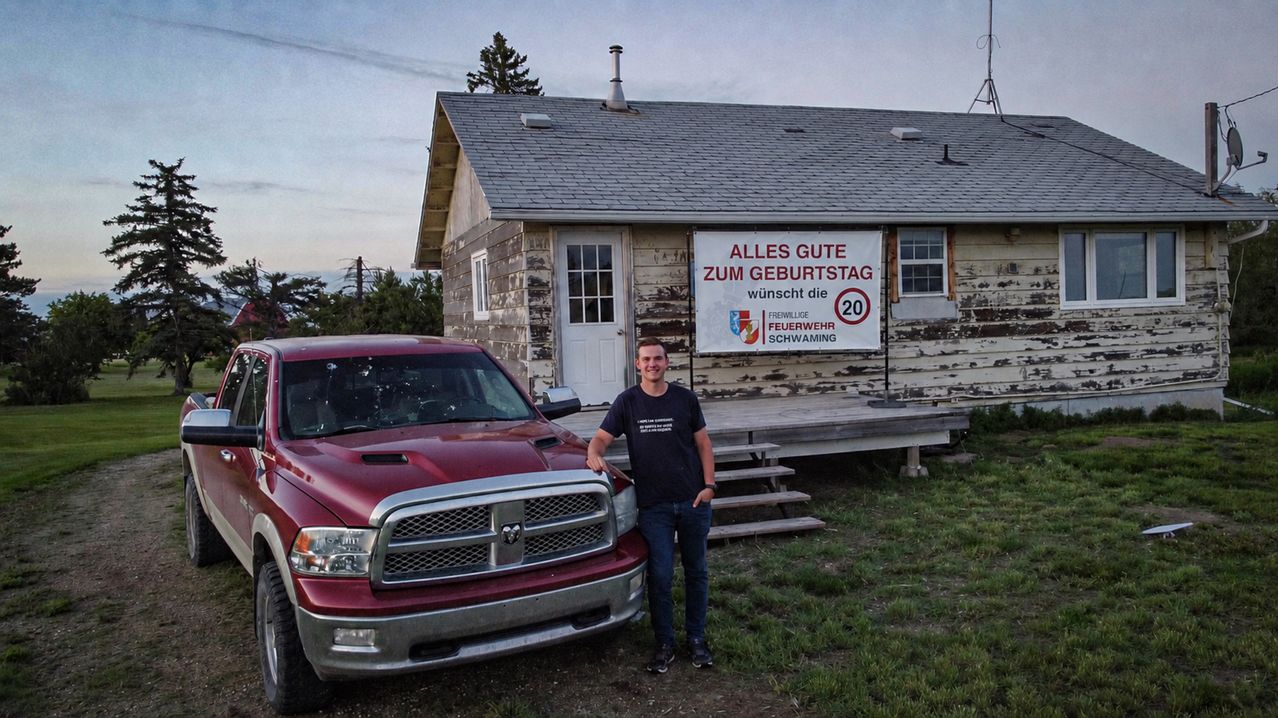 A man stands next to a red pickup truck in front of a small house with a sign that reads 'Alles Gute zum Geburtstag' and a speed limit of 20.