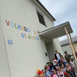 Eine Gruppe von Kindern und Erwachsenen versammelt sich vor dem Gebäude der St. Martin Volksschule, einige halten Blumen und ein Mann einen Topfpflanzen.