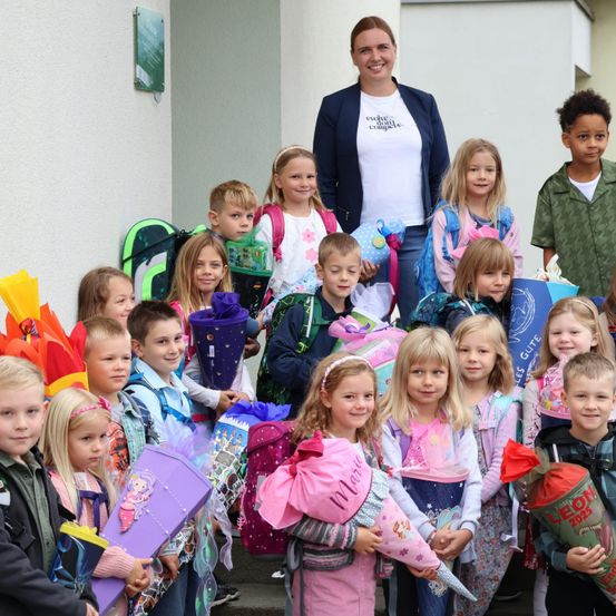 Eine Gruppe von Kindern und Erwachsenen steht lächelnd draußen, einige halten Geschenke. Eine Frau in einem weißen T-Shirt steht hinter den Kindern. Die Kinder tragen Rucksäcke und halten verschiedene Gegenstände.