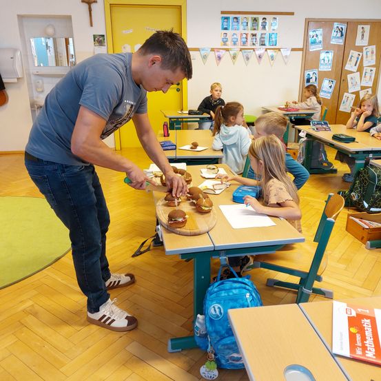 Ein Mann in einem blauen T-Shirt und Jeans bereitet Essen auf einem Schneidebrett in einem Klassenzimmer zu. Mehrere Kinder sitzen um ihn herum an Tischen.