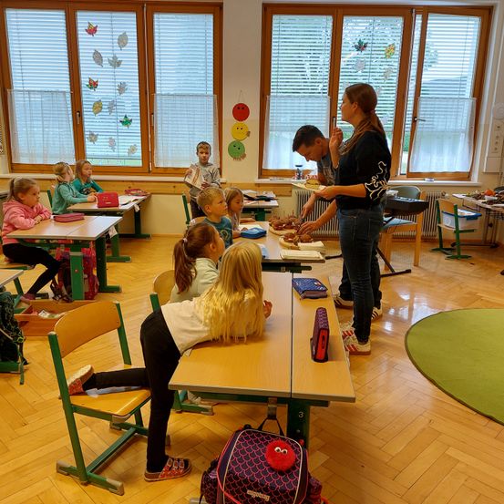 Ein Klassenzimmer mit Kindern, die an Tischen sitzen, eine Frau steht vorne und ein Mann verteilt Essen. Der Raum hat Fenster mit Jalousien und Dekorationen an der Wand.
