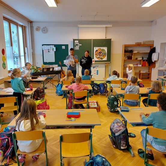 Klassenzimmer mit Kindern an Schreibtischen. Zwei Erwachsene stehen vorne, einer hält ein Geschenk. Bunte Poster und eine Uhr schmücken die Wände. Verschiedene Bücher, Taschen und Musikinstrumente sind verstreut.