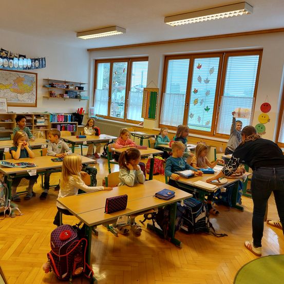 In einem Klassenzimmer sitzen mehrere Kinder an Schreibtischen. Ein Kind hebt die Hand, und eine Lehrerin kümmert sich um es. Auf den Schreibtischen befinden sich Rucksäcke und Bücher. Das Klassenzimmer hat große Fenster mit Jalousien und Dekorationen.