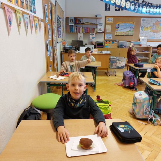 Ein Klassenzimmer mit Kindern an Schreibtischen, ein Junge vorne lächelt und hält ein Sandwich, mit Rucksäcken und verschiedenen Gegenständen in der Umgebung.